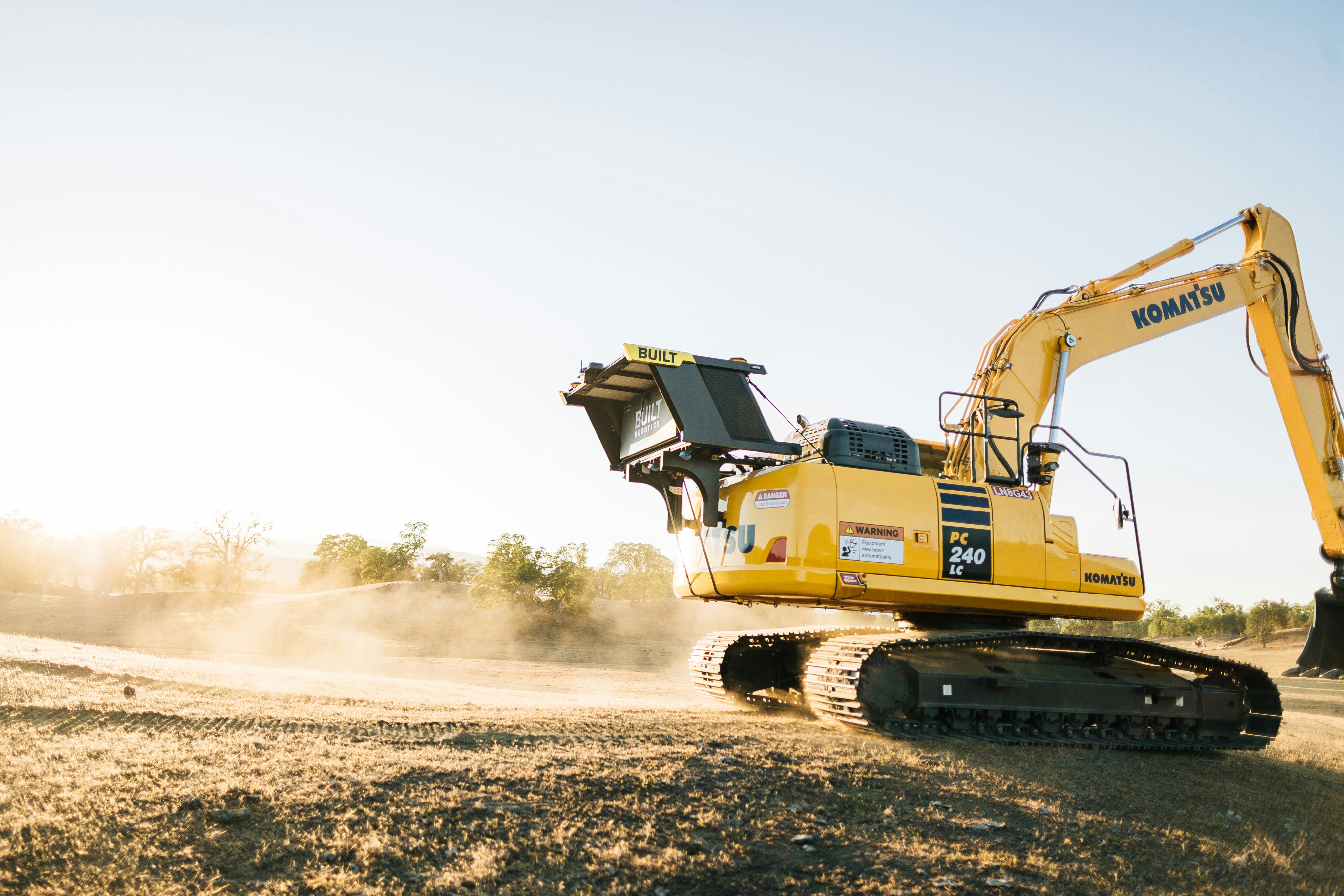 Komatsu PC240LC excavator grading a dry construction site at sunrise, tracked heavy equipment kicking up dust during earthmoving.