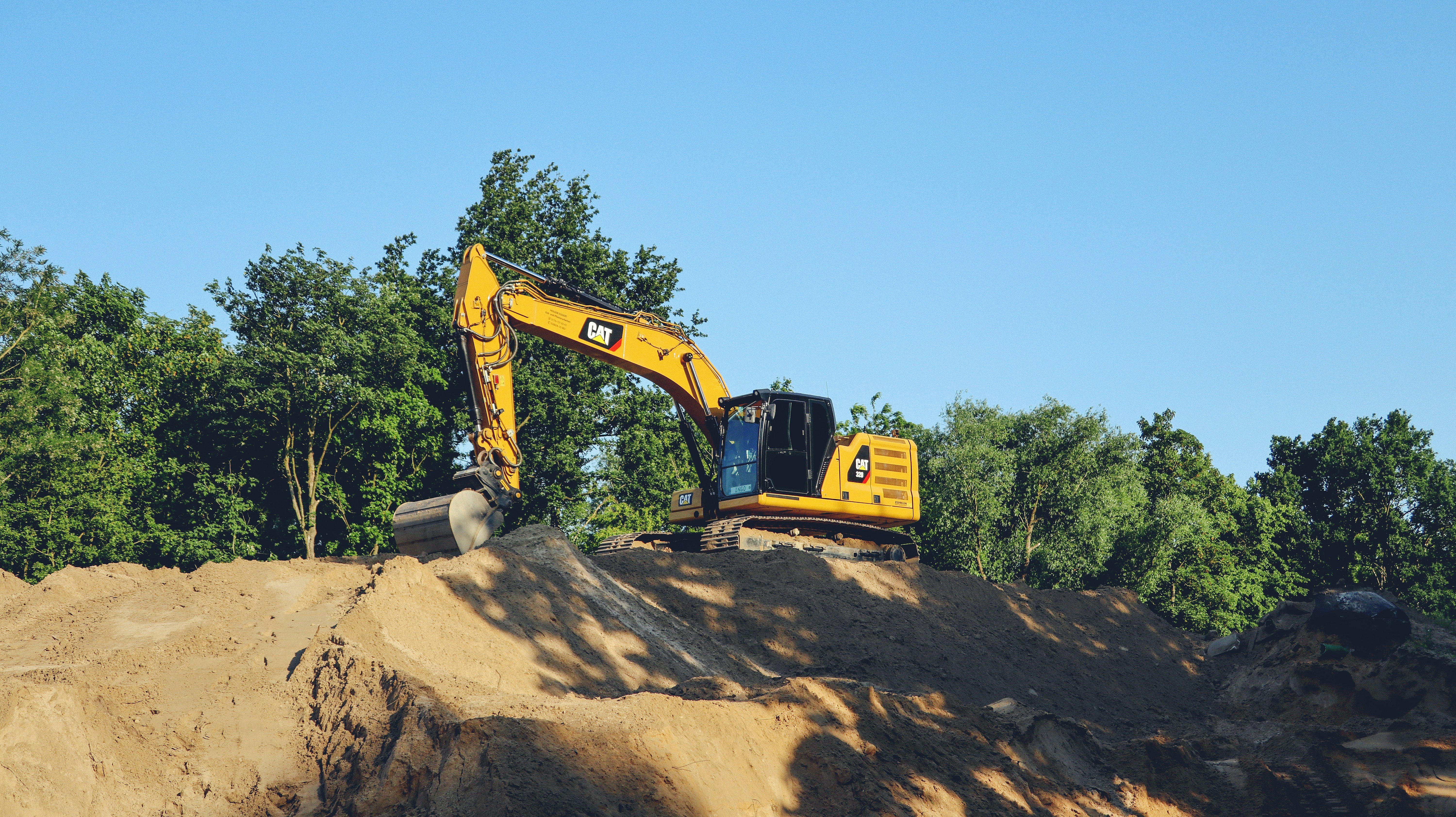 A yellow CAT excavator positioned on top of a large dirt mound or hill, with its bucket attachment visible. The machine is surrounded by green trees under a clear blue sky, appearing to be at a construction site.