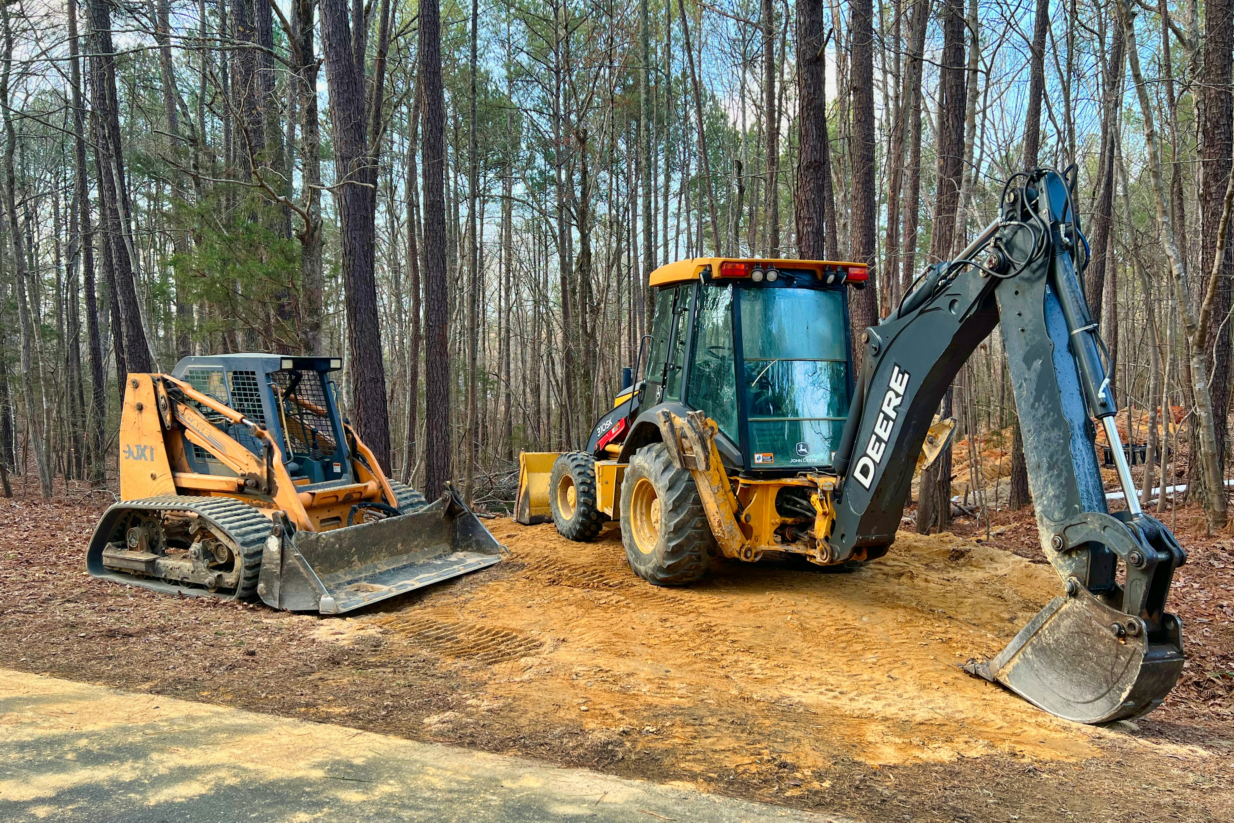 Compact track loader and John Deere backhoe parked in a forested work zone, demonstrating the versatility of small and mid-sized construction equipment in tight or wooded environments.