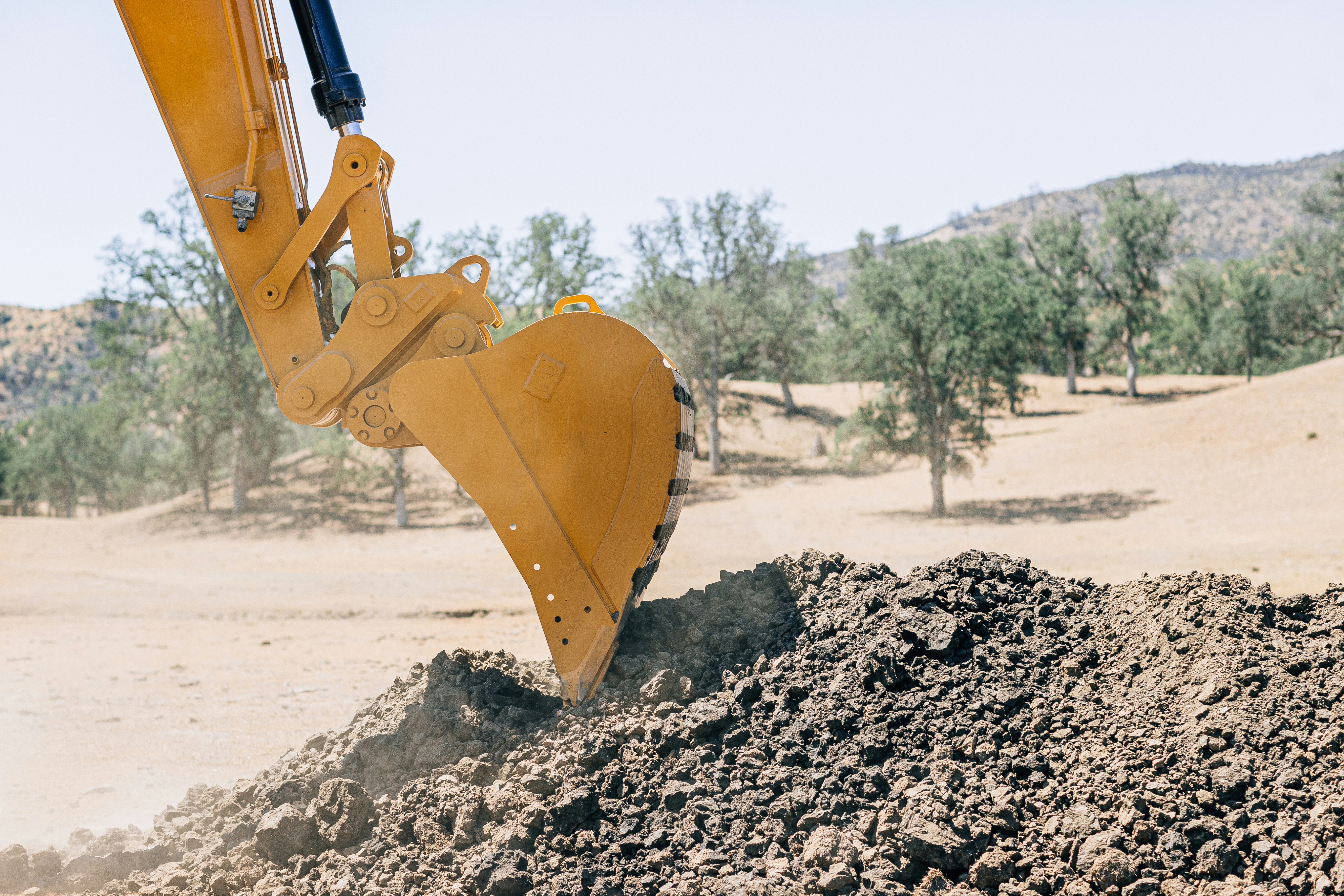Close-up view of a yellow excavator bucket digging into dark soil or dirt, with a desert-like landscape visible in the background featuring sparse vegetation and rolling hills under an overcast sky.