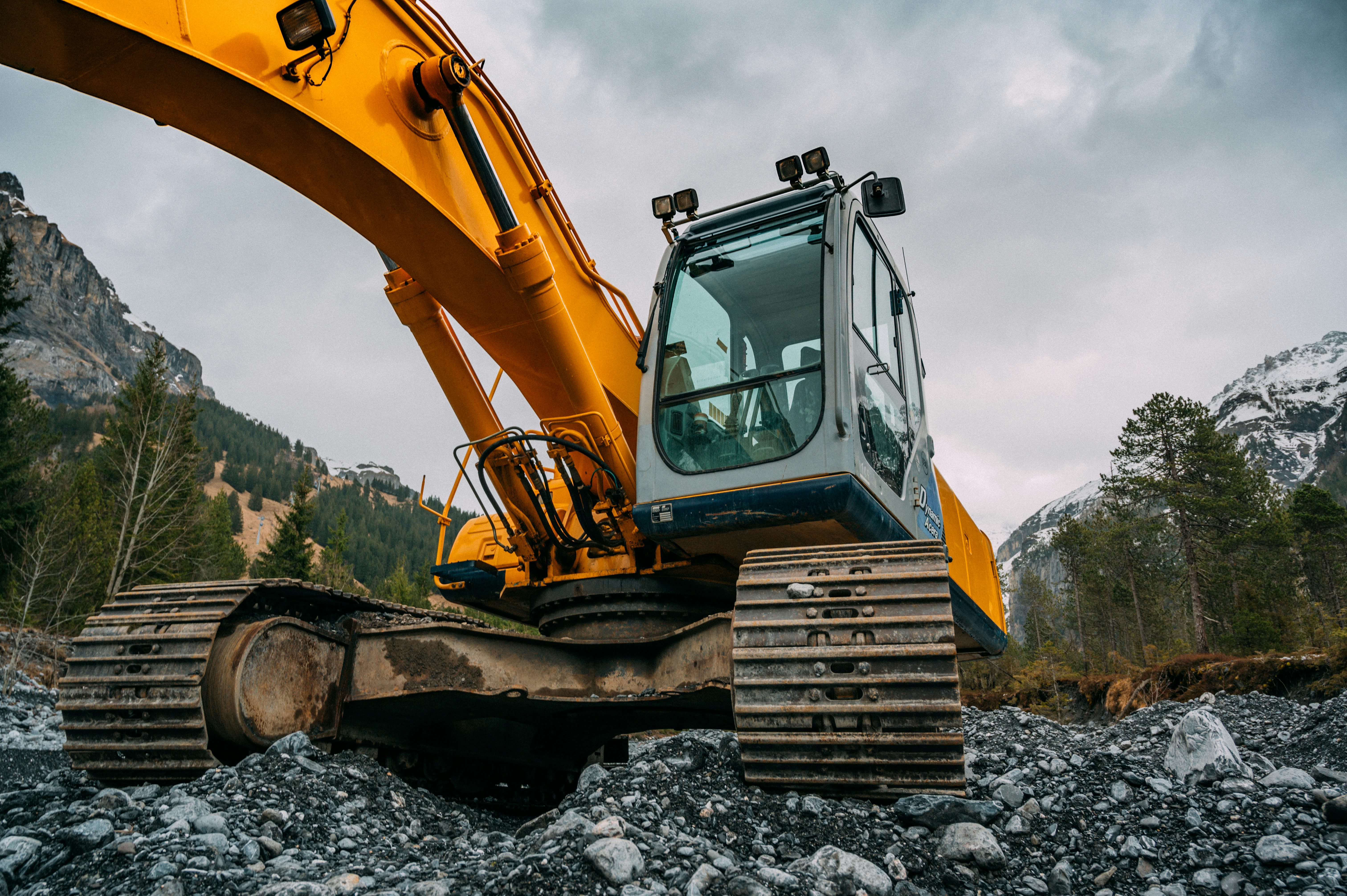 A yellow and white excavator with rubber tracks positioned on rocky, gravelly terrain in a mountainous valley.