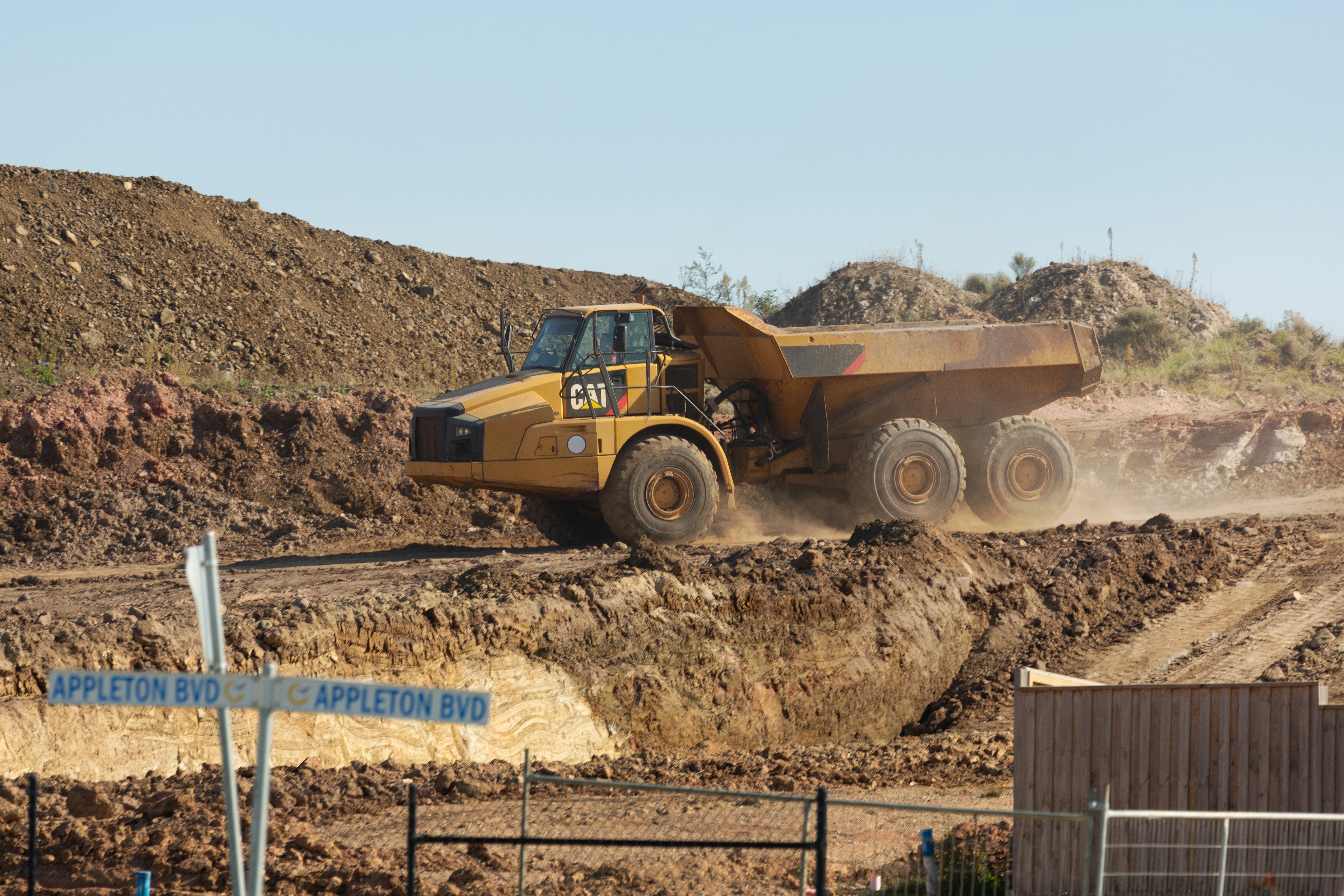 CAT articulated dump truck hauling dirt across an active construction site near Appleton Boulevard, with dust trailing from the wheels.