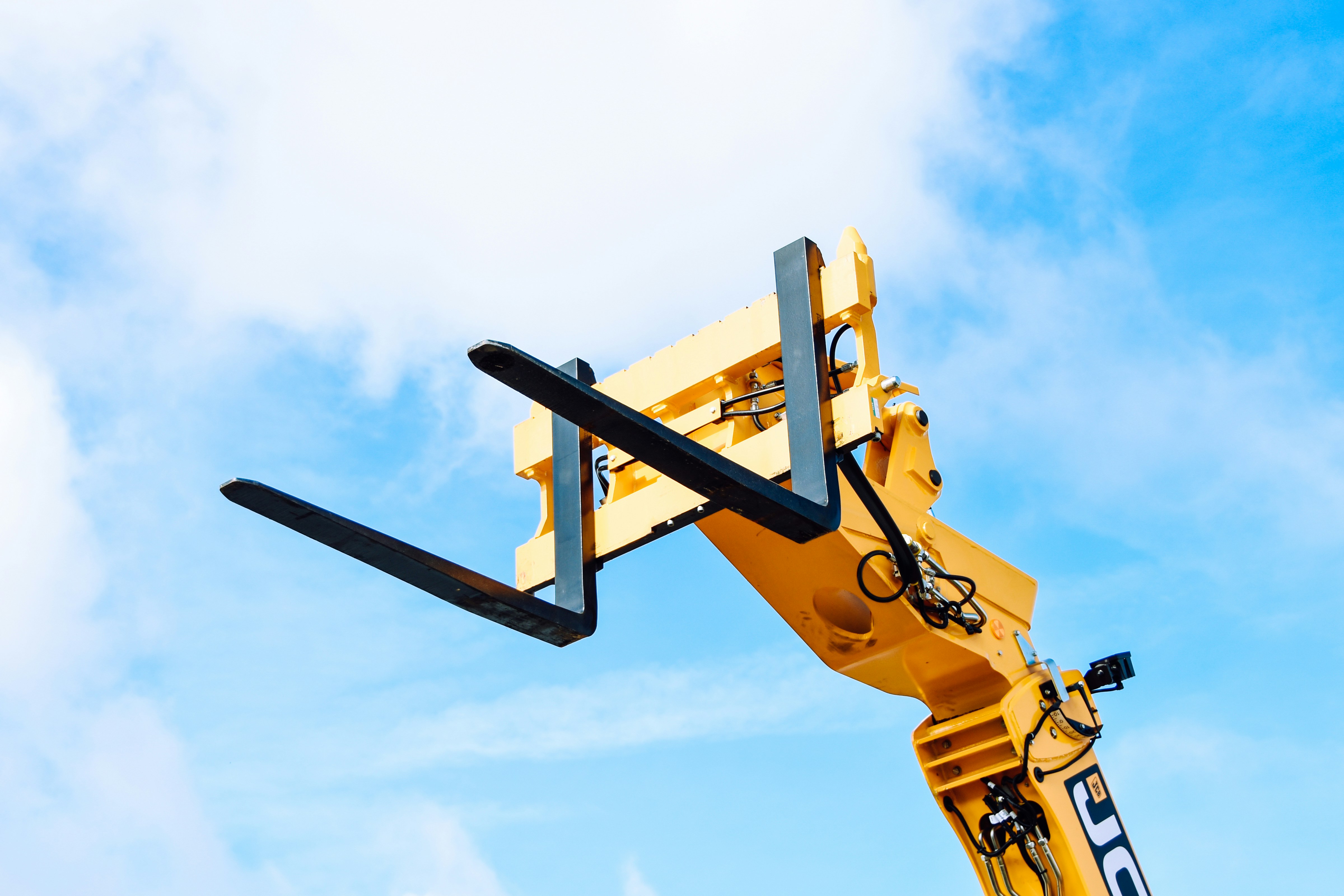 JCB telehandler boom with pallet-fork attachment raised against a blue sky, material-handling equipment for construction sites.