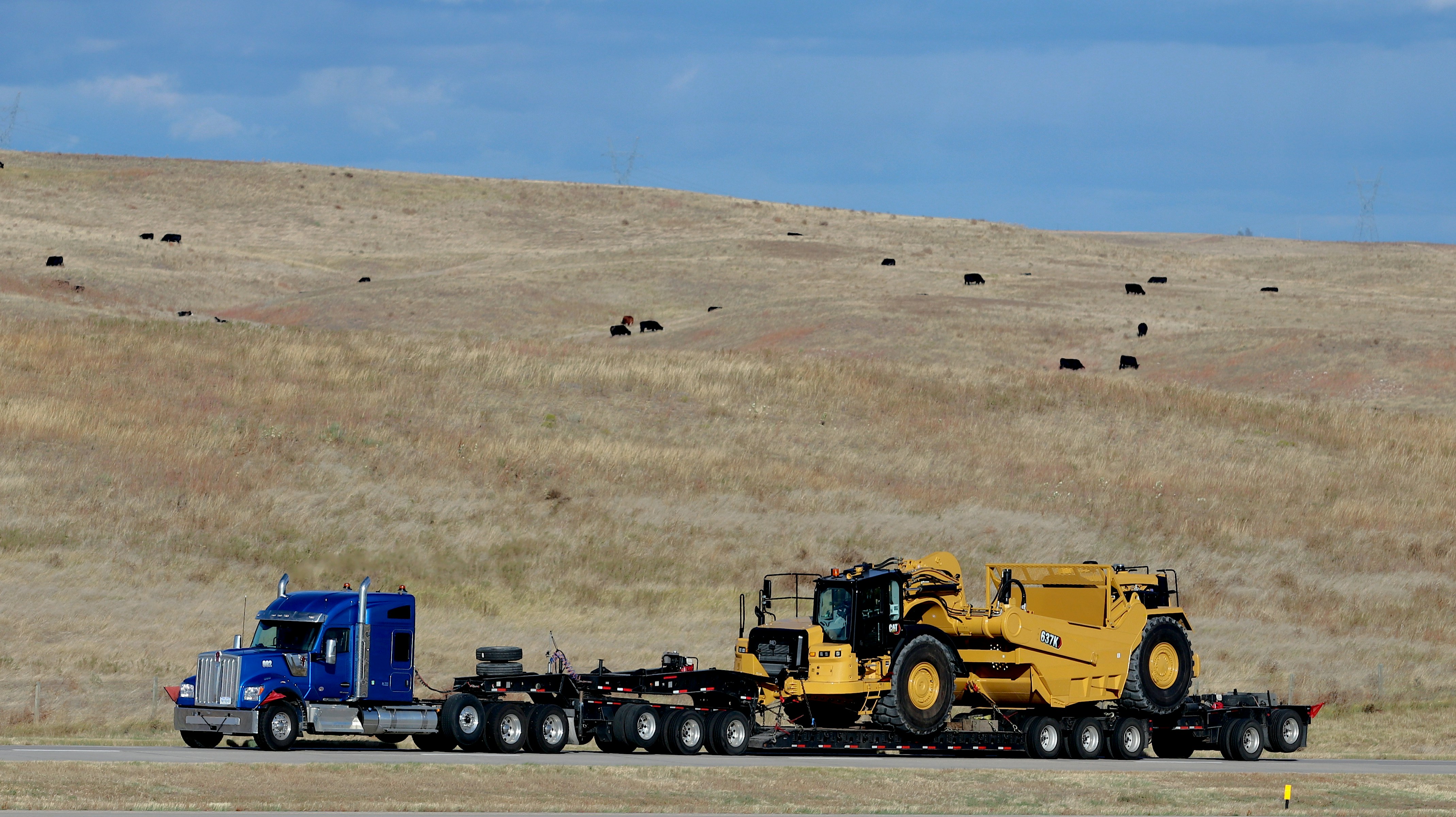 Heavy-duty semi-truck hauling a Caterpillar 637K scraper across rural terrain, showcasing long-distance heavy equipment transport by lowboy trailer.