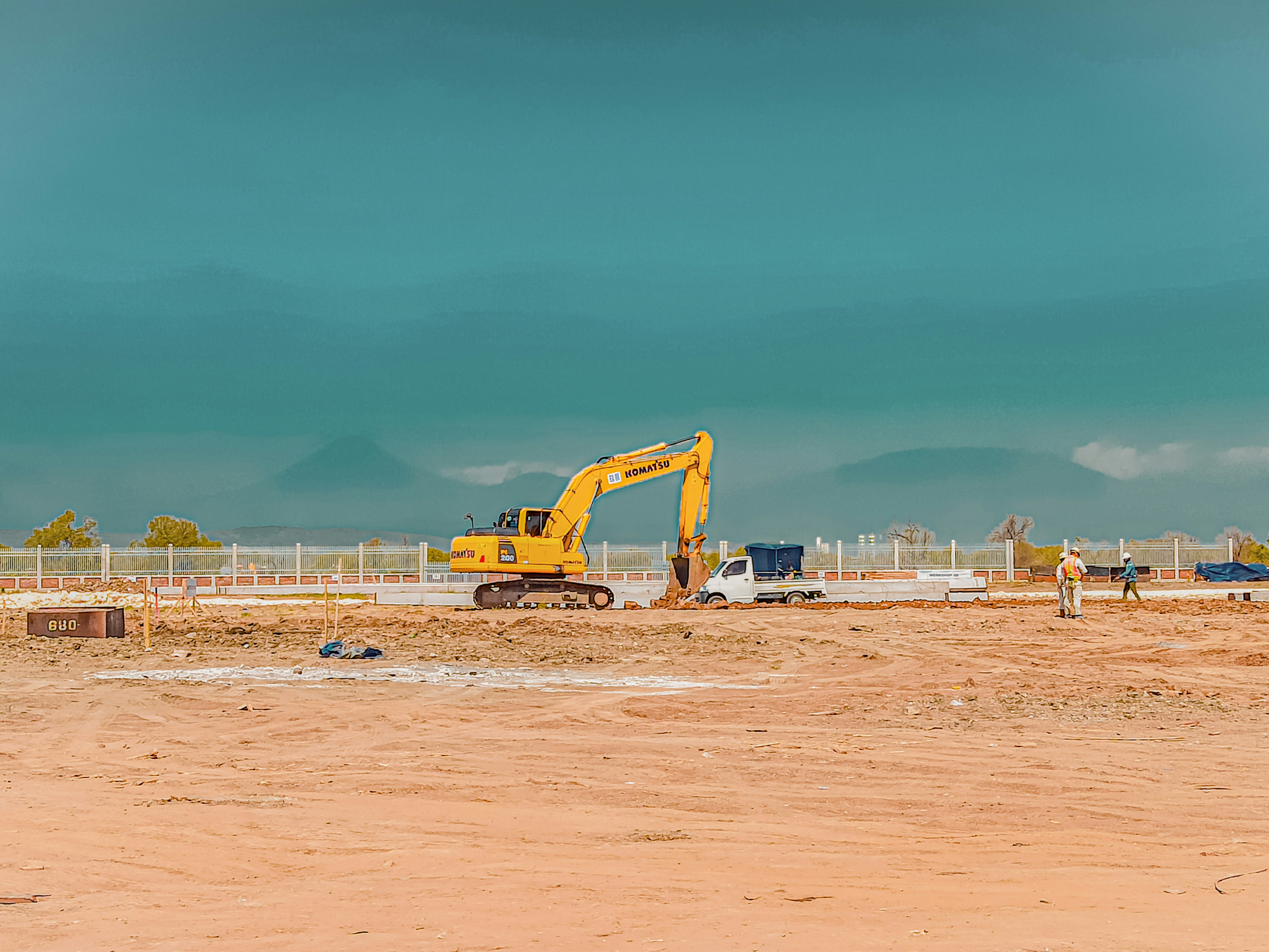 Komatsu hydraulic excavator digging on a commercial construction site with workers nearby under a clear turquoise sky.
