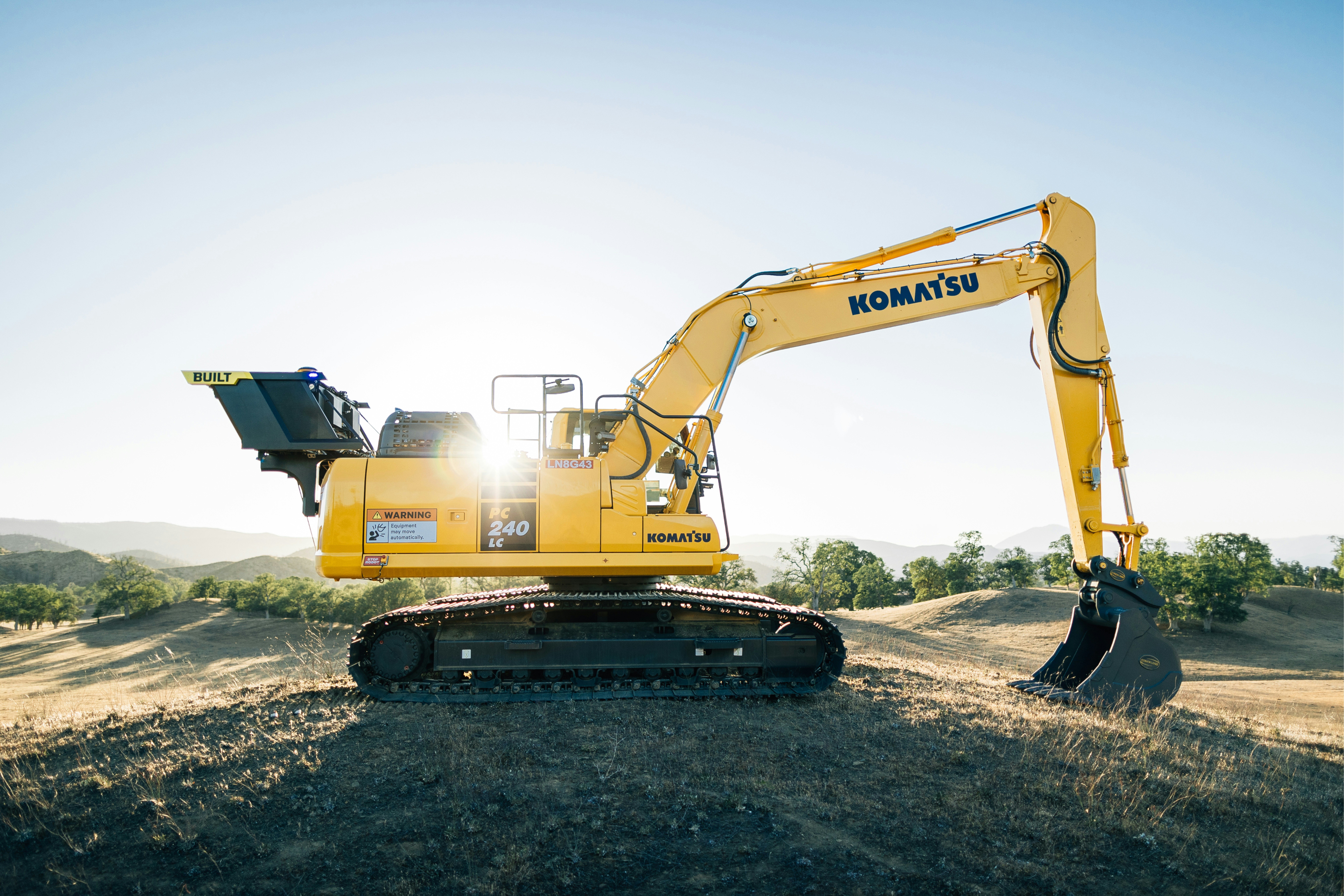 Side profile of Komatsu PC240LC excavator on a hillside with sun flare, tracked excavator for precision digging and grading.