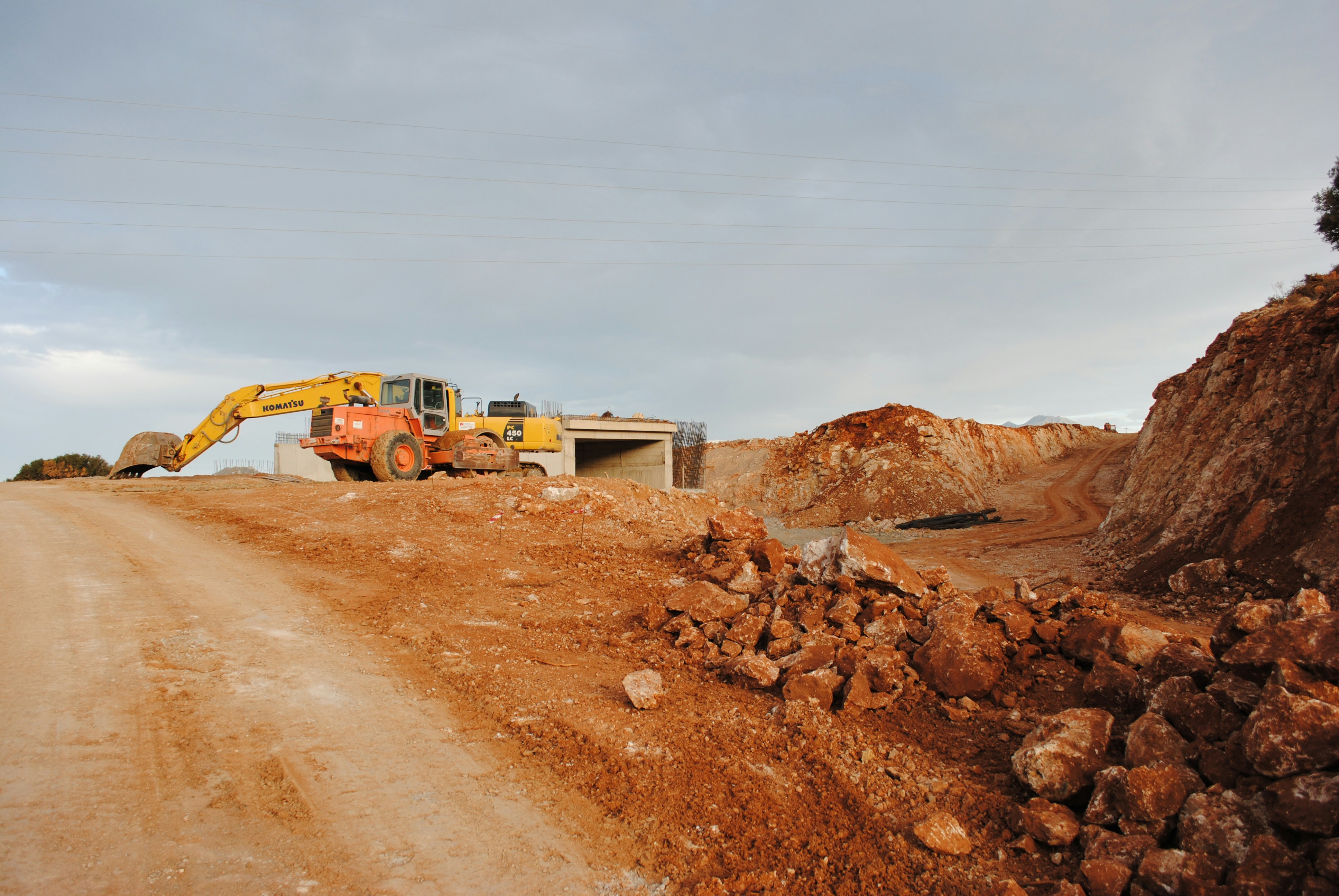 Komatsu PC450 excavator working on a road construction project, positioned beside a partially built concrete structure and surrounded by rocky terrain.