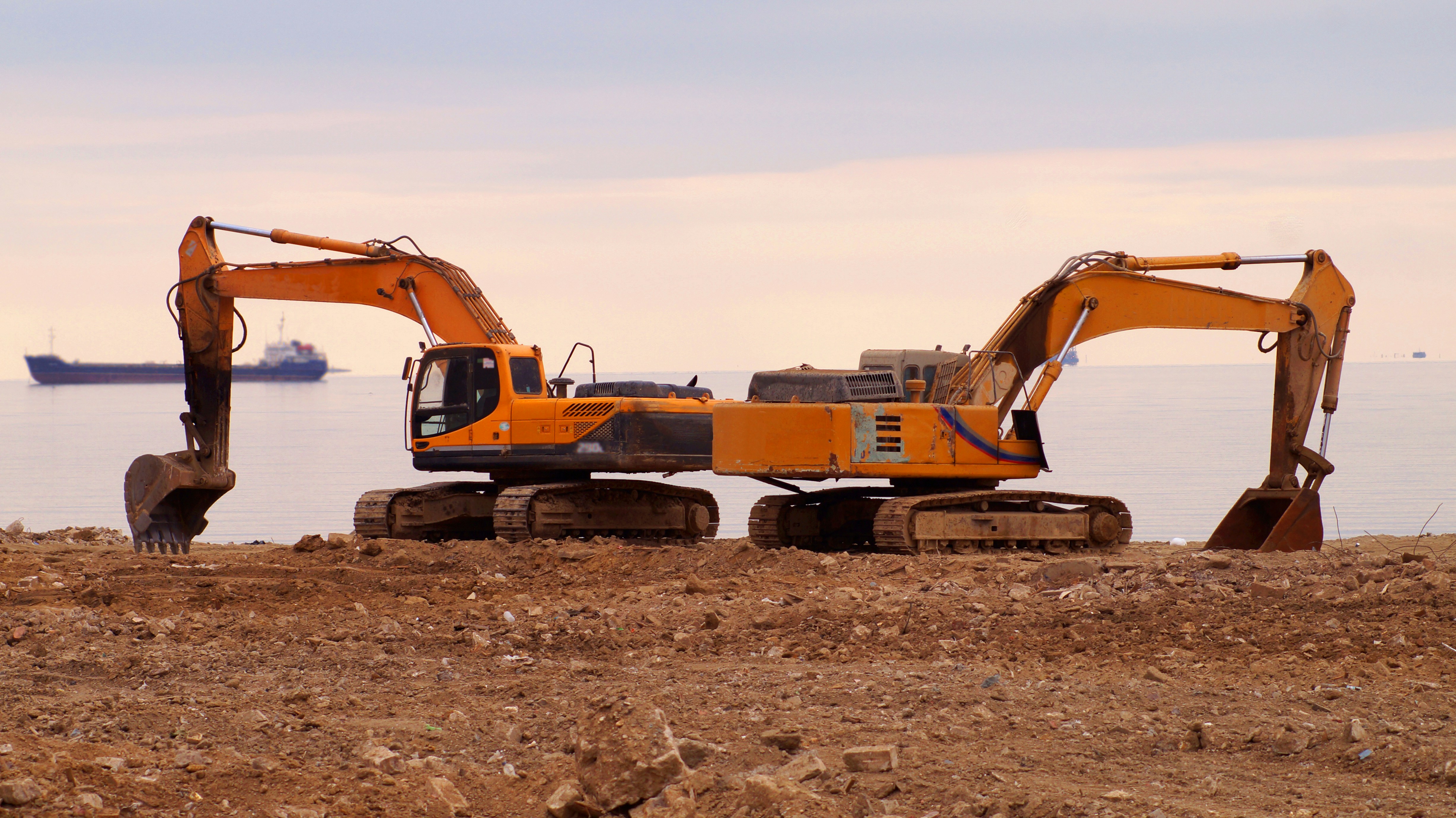Two tracked excavators positioned on a coastal job site with ships in the background, representing durable construction equipment used for marine and shoreline projects.